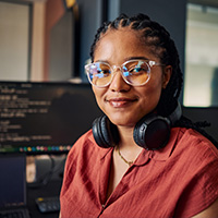 A professional-looking woman with a warm smile, wearing glasses and headphones, in a tech-oriented environment.